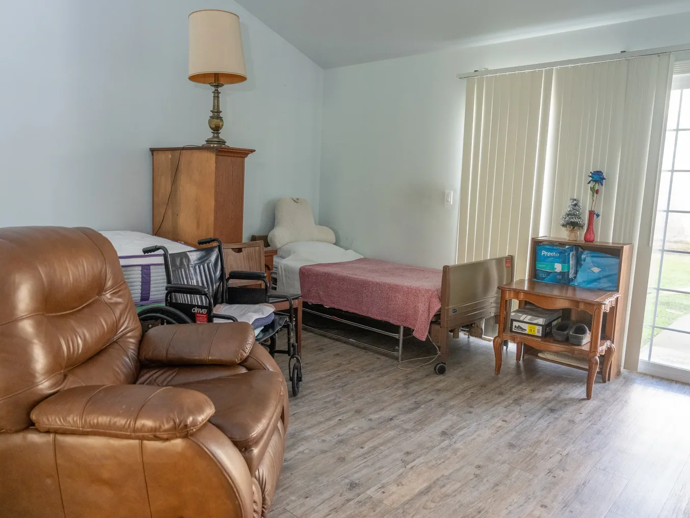 Spacious accessible bedroom featuring hospital bed, wheelchair, comfortable recliner and sliding glass door with natural light